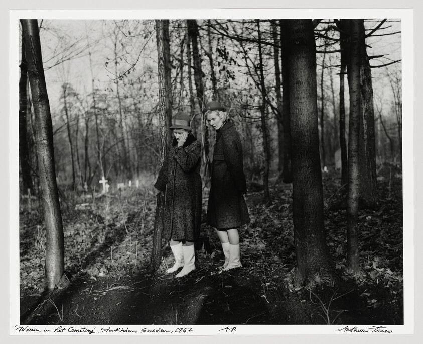 Two women in coats stand in a wooded cemetery, one covering her face.