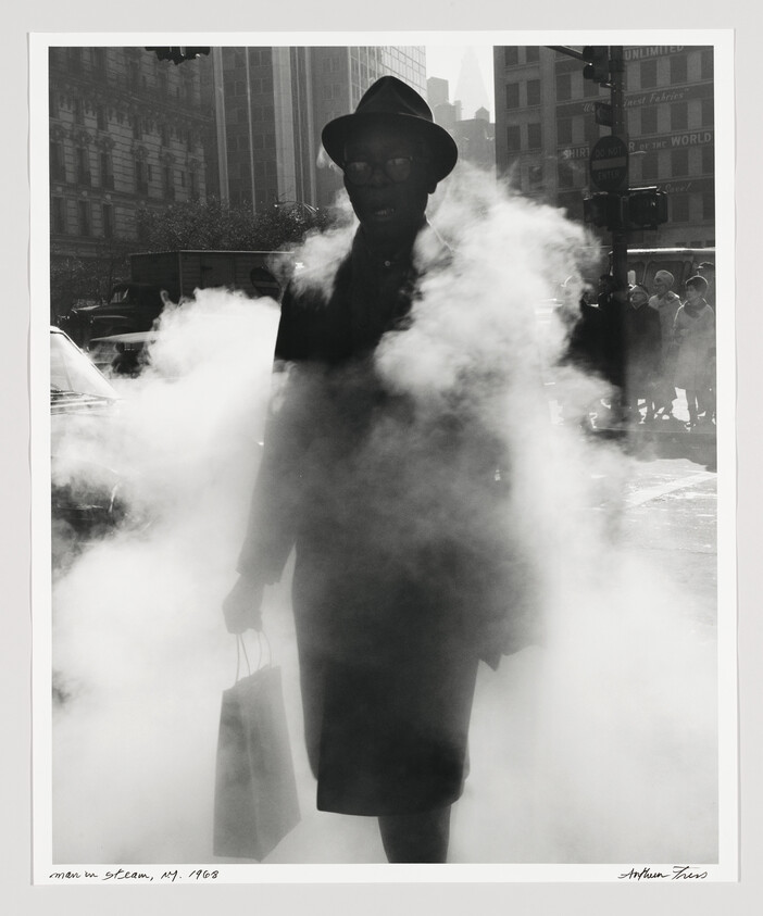 A person in a hat walks through thick street steam while holding a shopping bag.