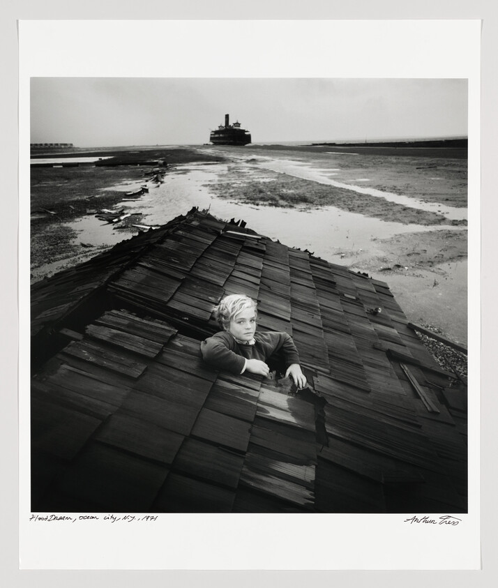 Young boy climbs out of a broken rooftop on a muddy shoreline with a ship in the distance.