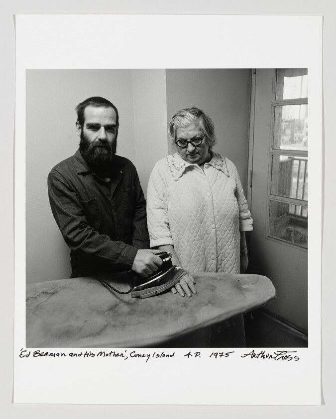 A bearded man presses an iron near an older woman's hand on an ironing board.