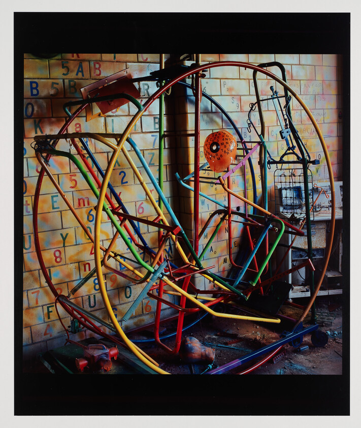 Colorful tangled circular metal playground equipment leaning against a tiled wall with letters and numbers.