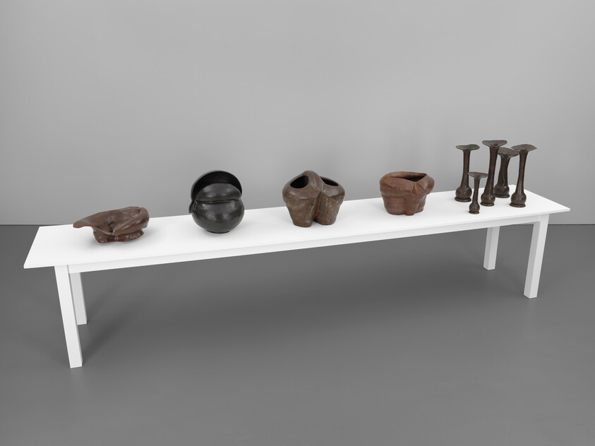 A long white table displays six organic-shaped brown and black ceramic vessels and tall candlestick sculptures.