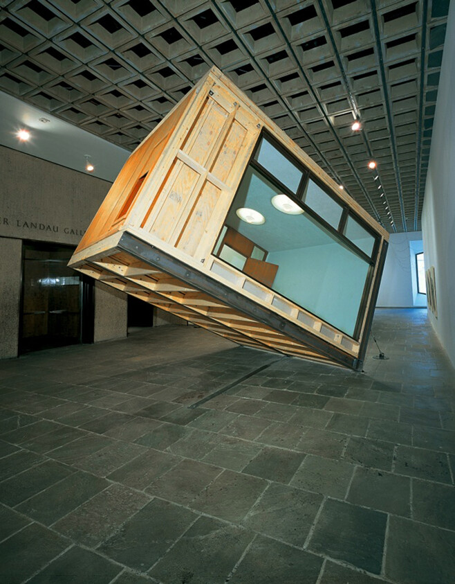 A large wooden room module tilted and seemingly balancing on one corner inside a gallery.