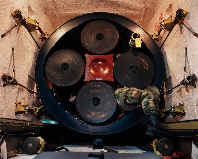 A soldier in camouflage leans into a circular rack of large missile canisters inside a cramped aircraft bay.