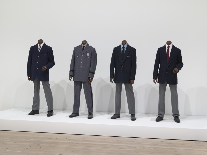 Four headless mannequins wear different dark blazers and gray trousers standing on a white platform.