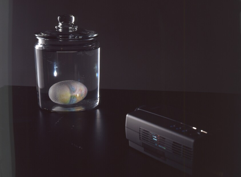 A glass jar on a table contains a colorful submerged egg while a small projector sits nearby.