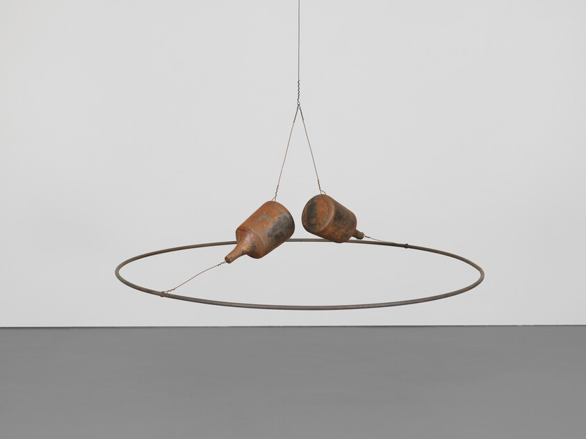 Two rusted weights hang from chains above a suspended metal ring in an empty gallery.