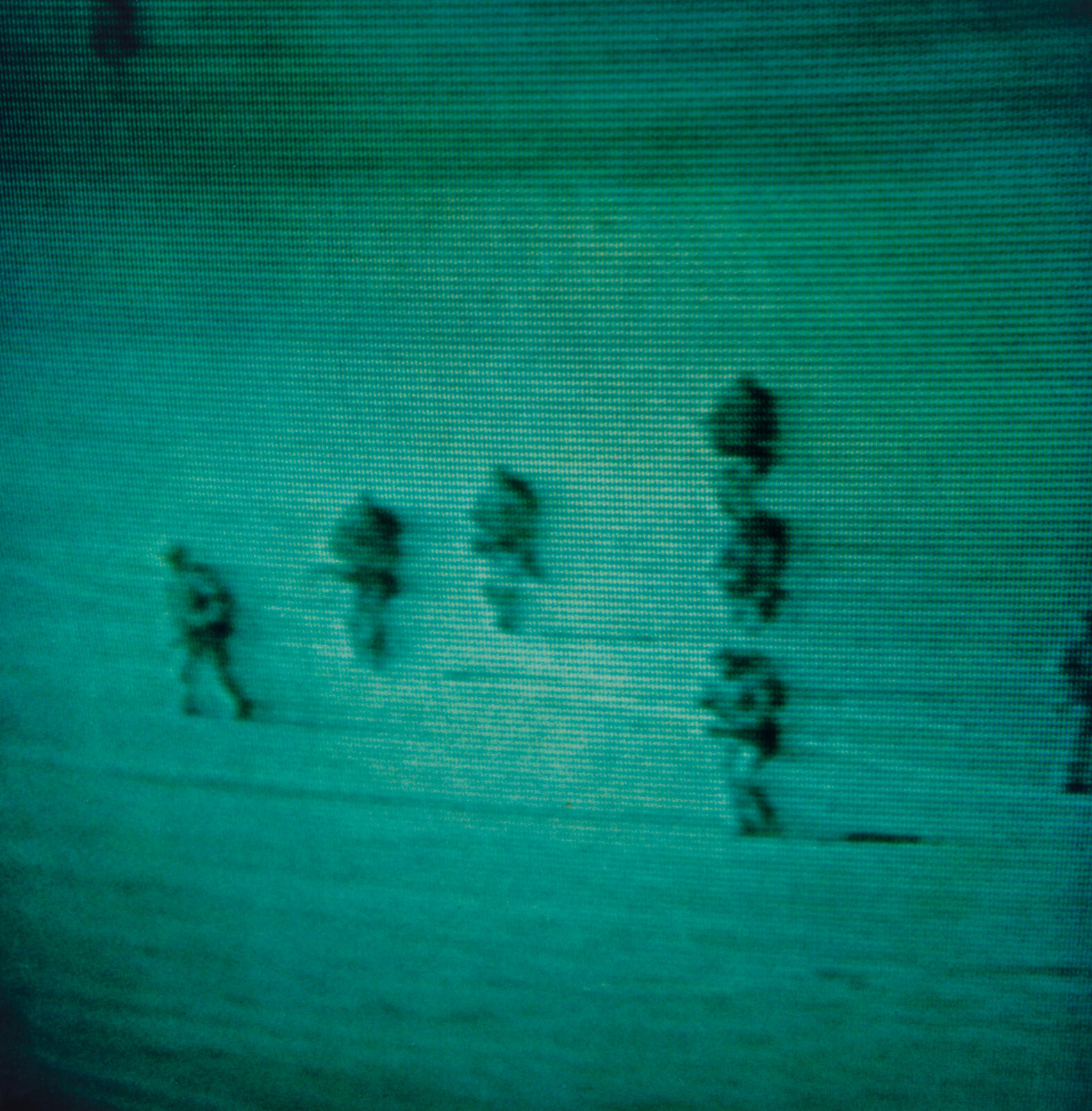 Several people wearing backpacks walk in a line across a snowy field under green night vision.