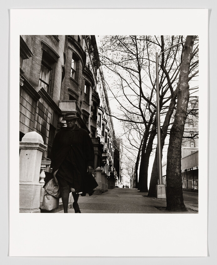 A black and white photograph capturing a city street lined with bare trees and classic buildings, with a blurred figure in the foreground suggesting motion, possibly a person walking by quickly.