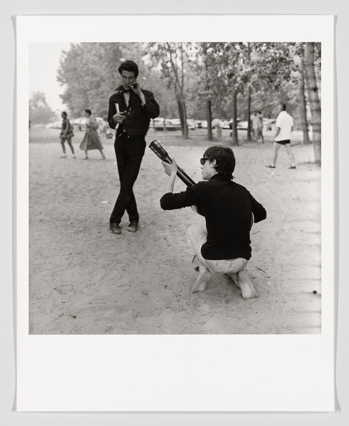 A young man kneels on sand playing a guitar while another man stands smoking nearby.