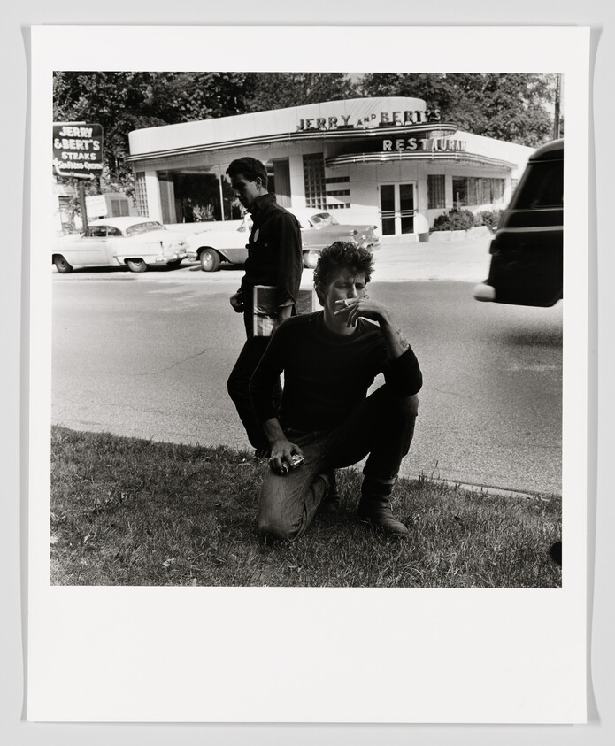 Young man kneels smoking by the roadside while another man stands nearby.
