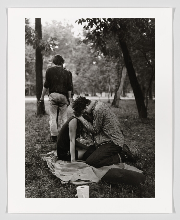 A couple kneels on a blanket in a park, embracing and touching foreheads while someone walks away.