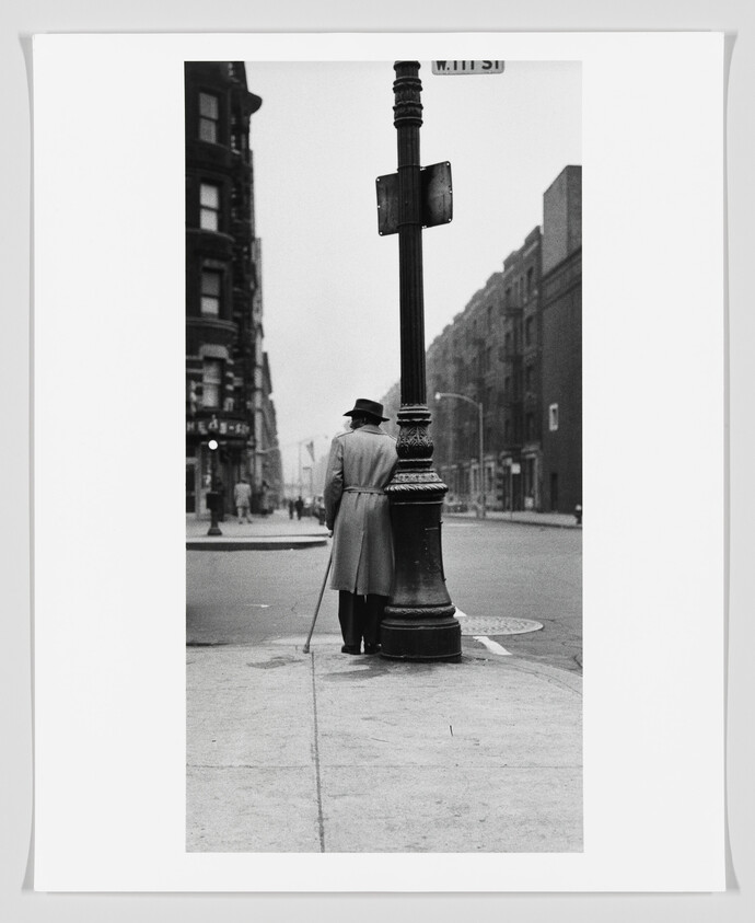 Man in a coat and hat leans on a streetlamp while holding a cane.