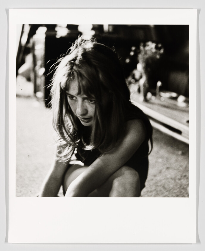 A young girl crouches on the ground looking down with a thoughtful, somber expression.