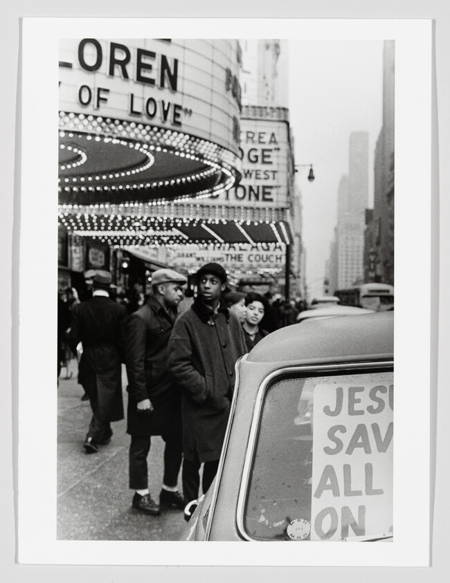 Young man in coat stands under a lit theater marquee beside a car with "Jesus saves" sign.