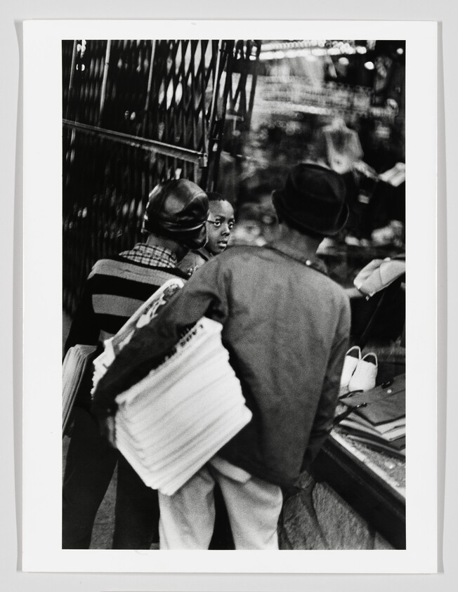 Three street vendors stand at a shop window, one carrying a large stack of newspapers.