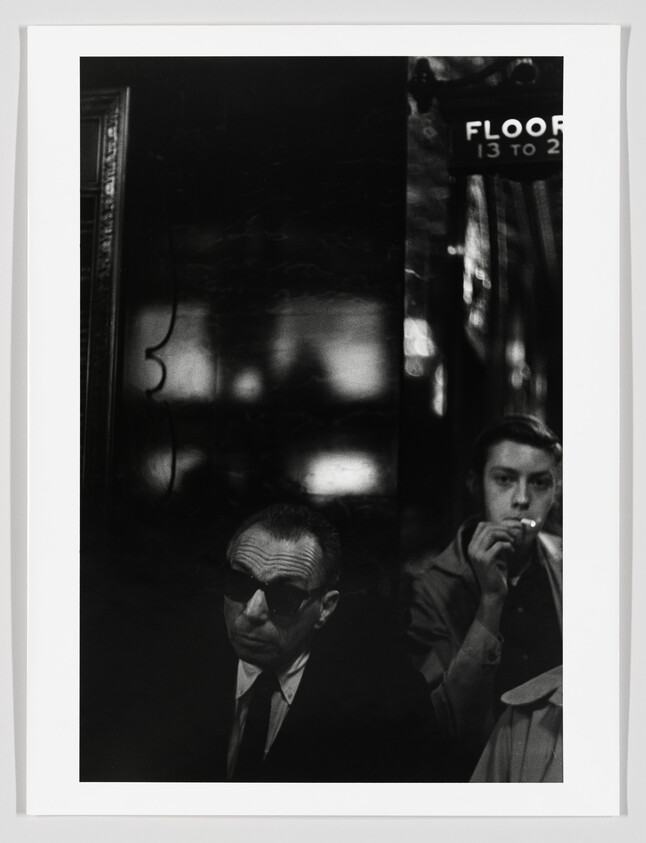 A man in sunglasses sits in shadow while another man beside him smokes a cigarette.