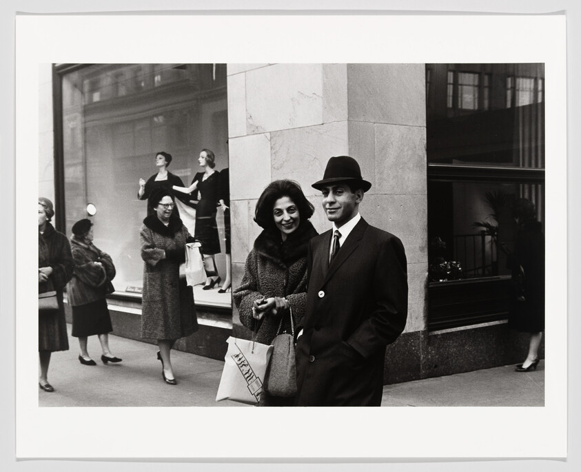 A smiling well-dressed couple stands on a city sidewalk outside a department store, the woman holding a shopping bag.