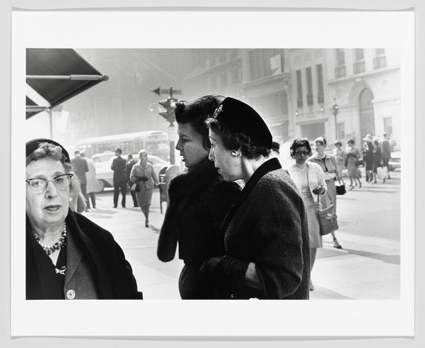 Three women in coats stand and walk on a busy city sidewalk, older woman looking toward camera.