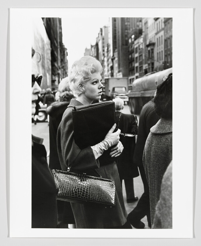 A blonde woman stands in a city crowd holding a large folder and handbag.