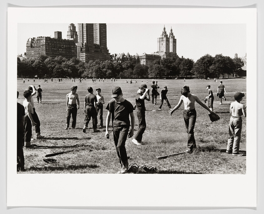 Boys playing informal baseball in a large park field with city skyline and apartment buildings behind.