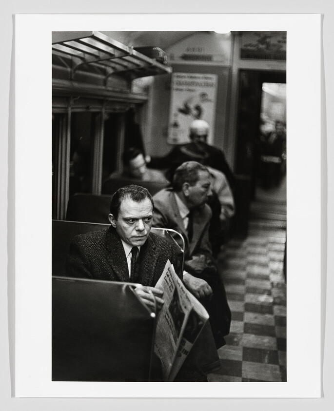 A serious man sits on a subway car holding a folded newspaper while other passengers sit.