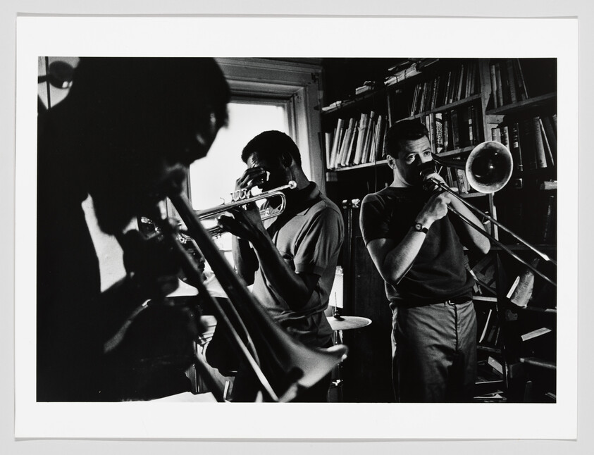 Three musicians play brass instruments together in a cramped room lined with bookshelves.