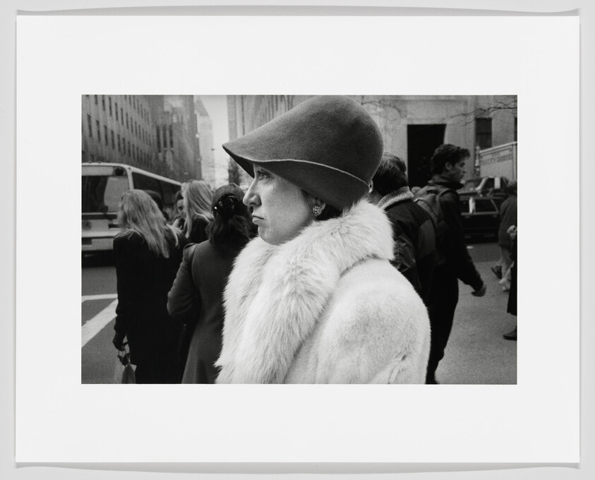 Woman in a fur coat and cloche hat stands on a busy city sidewalk among pedestrians.