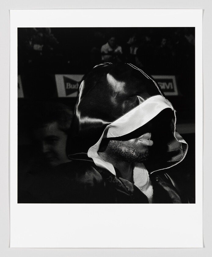 A black and white photograph capturing a close-up of a person wearing a hooded robe, with the hood casting a shadow over their face, leaving only the lower half of the face visible. The background is dimly lit with indistinct figures, suggesting the setting may be an event or a public gathering.