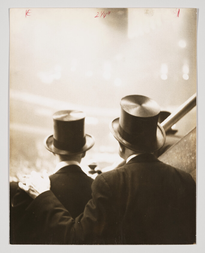 Two men in top hats and suits stand close together watching an event, one hand on shoulder.