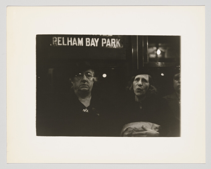 Two women sit in a dim subway car beneath a sign reading Pelham Bay Park.