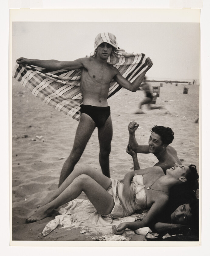 Young man spreads a striped towel like wings while friends laugh and relax on a sandy beach.