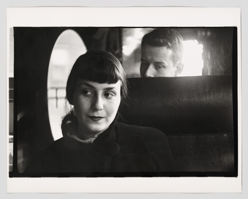 Young woman sits on a train seat looking to the side while a man watches from behind.