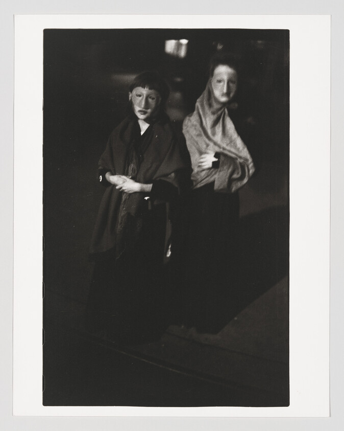 Two masked figures in dark cloaks stand close together with folded arms under dim lighting.