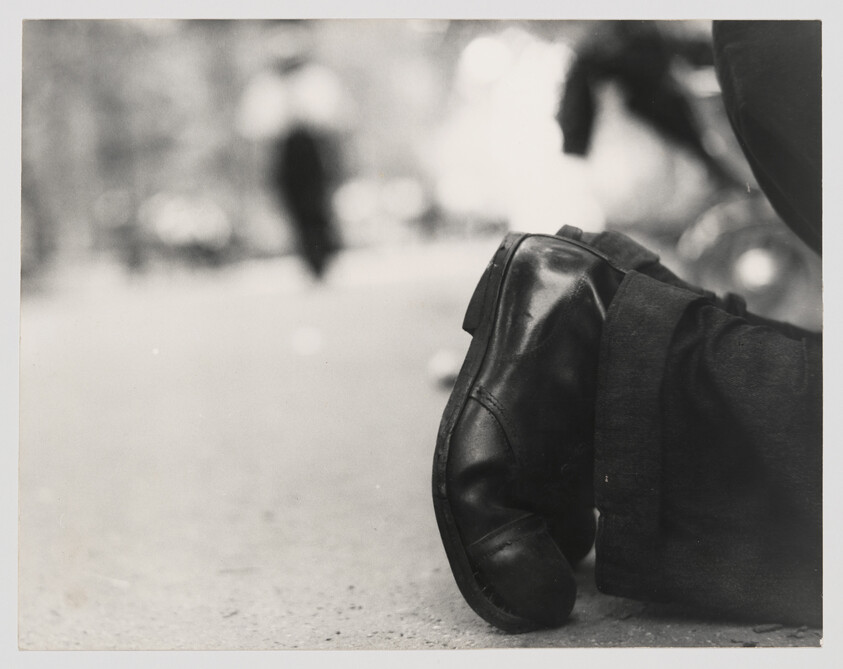Worn black dress shoes resting on crossed legs on a city sidewalk with blurred pedestrians.