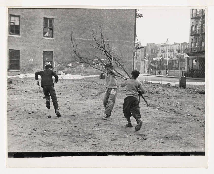 Three boys play in a dirt lot, with one carrying a large tree branch while others run nearby.