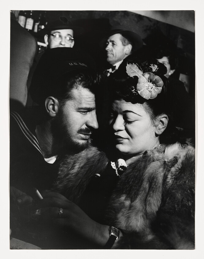A U.S. Navy sailor leans close to a woman wearing a fur coat and a floral hat.