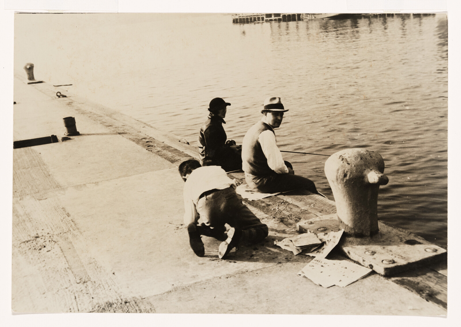 Three people sit and kneel on a dock while fishing by the water.