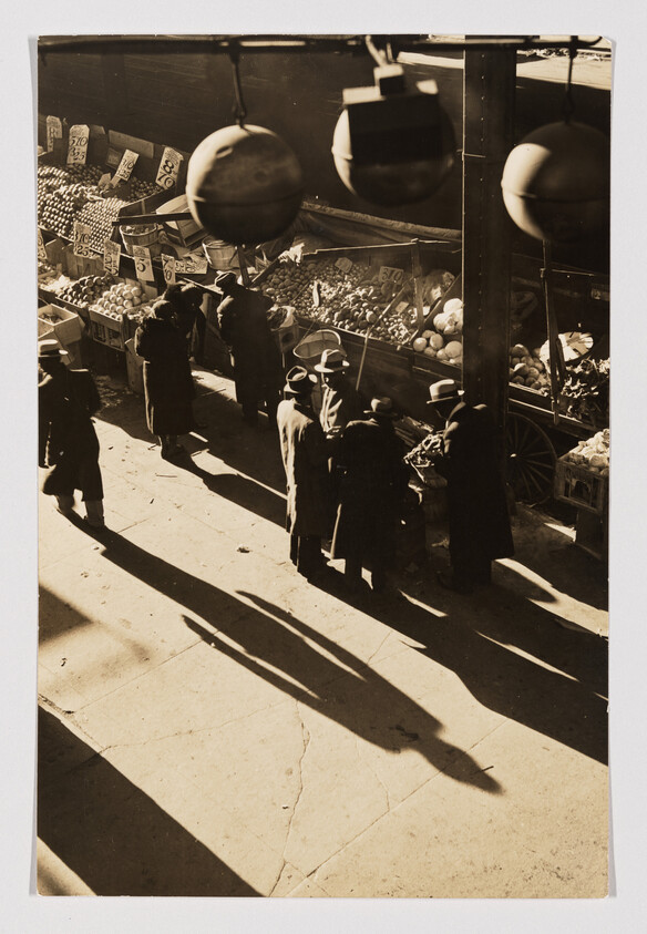 Shoppers in long coats gather at a street fruit and vegetable stall, casting long shadows.
