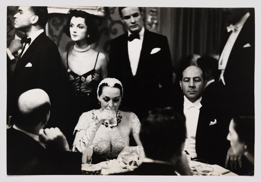 Woman in embellished dress drinks from a glass while formally dressed guests sit and stand around her.