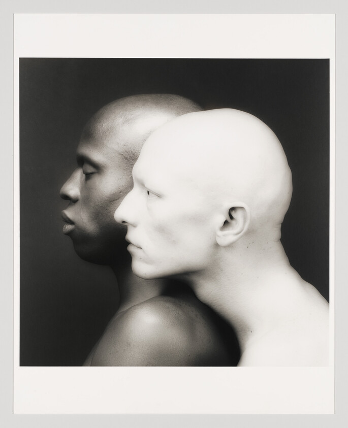Two men with contrasting skin tones stand closely side by side, profiles aligned and facing left.