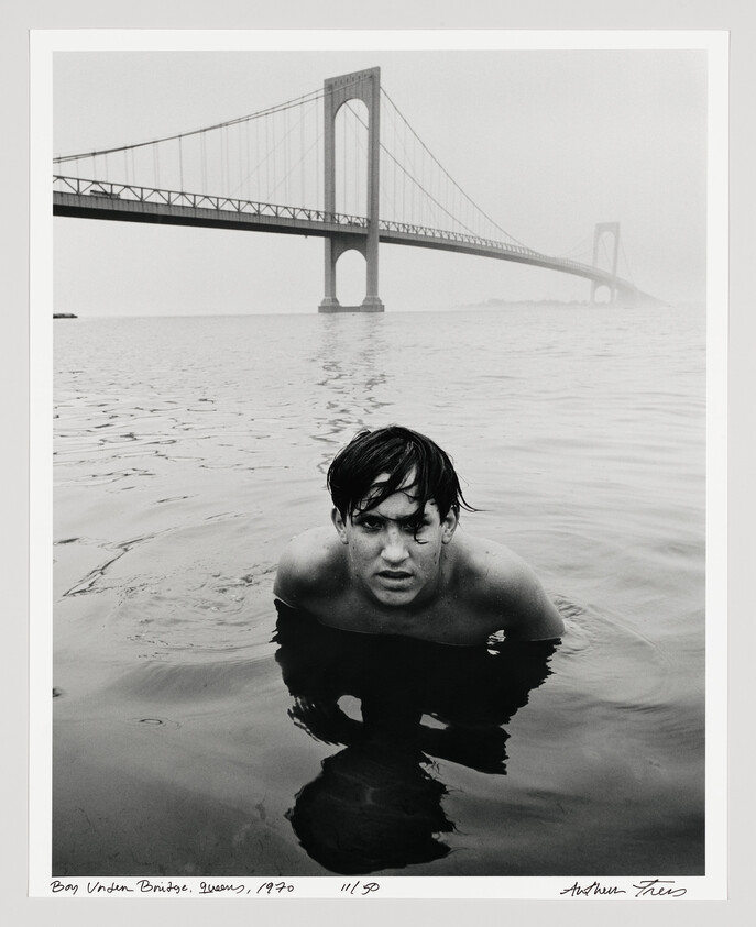 Teenage boy swims chest-deep in calm water with a large suspension bridge in the background.