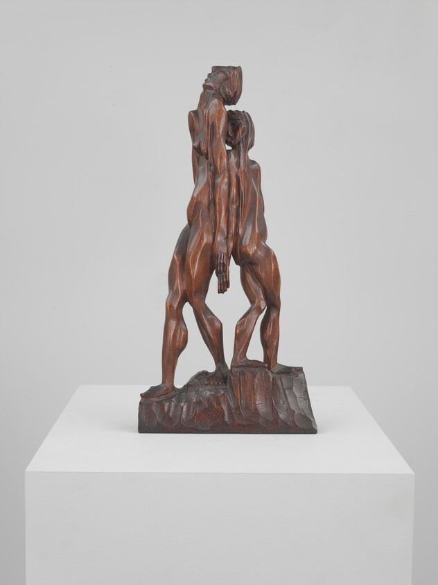 Two tall carved wooden figures leaning together on a rough base, one figure supporting the other.