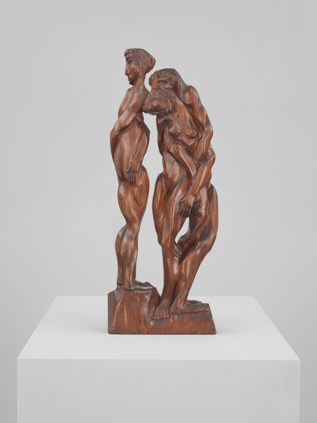 Two carved wooden human figures stand closely intertwined on a small pedestal, one leaning toward the other.