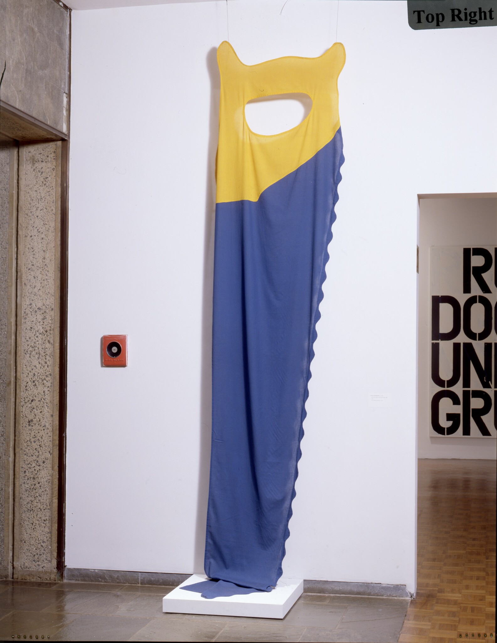 Tall hanging textile sculpture with yellow top and long blue draped panel suspended above a white base.