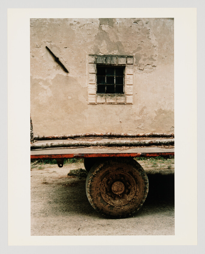 Rusty wagon wheel under a flatbed loaded with logs against a weathered plaster wall.
