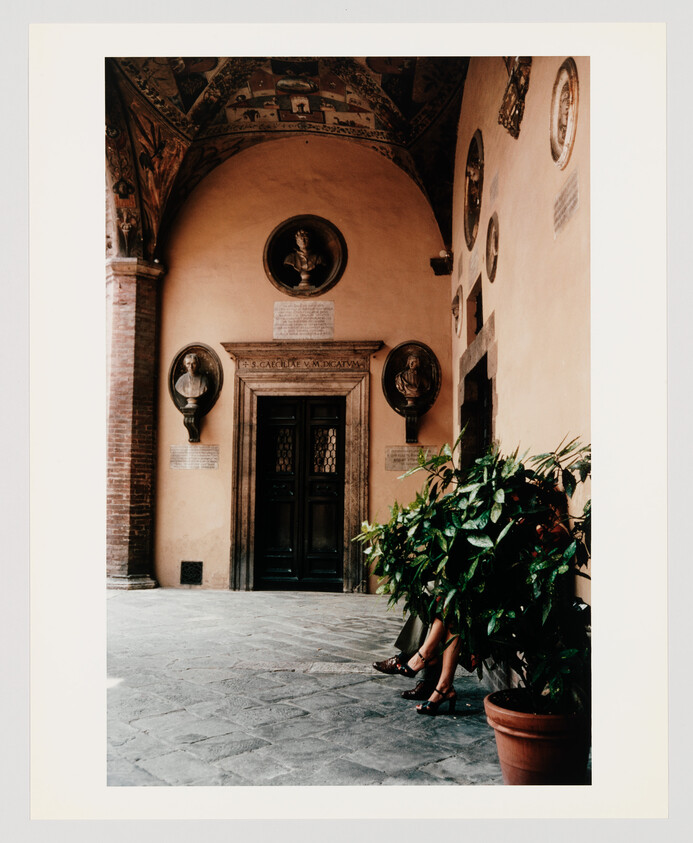 Ornate arched courtyard entrance with two people sitting behind a potted plant, their legs visible.