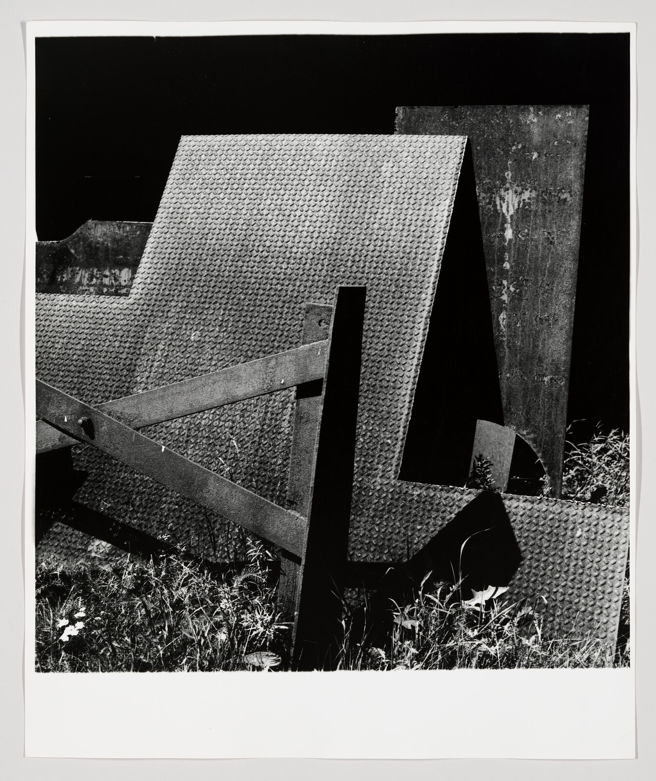 Rusty textured metal panels and wooden supports leaning amid tall grass and weeds.