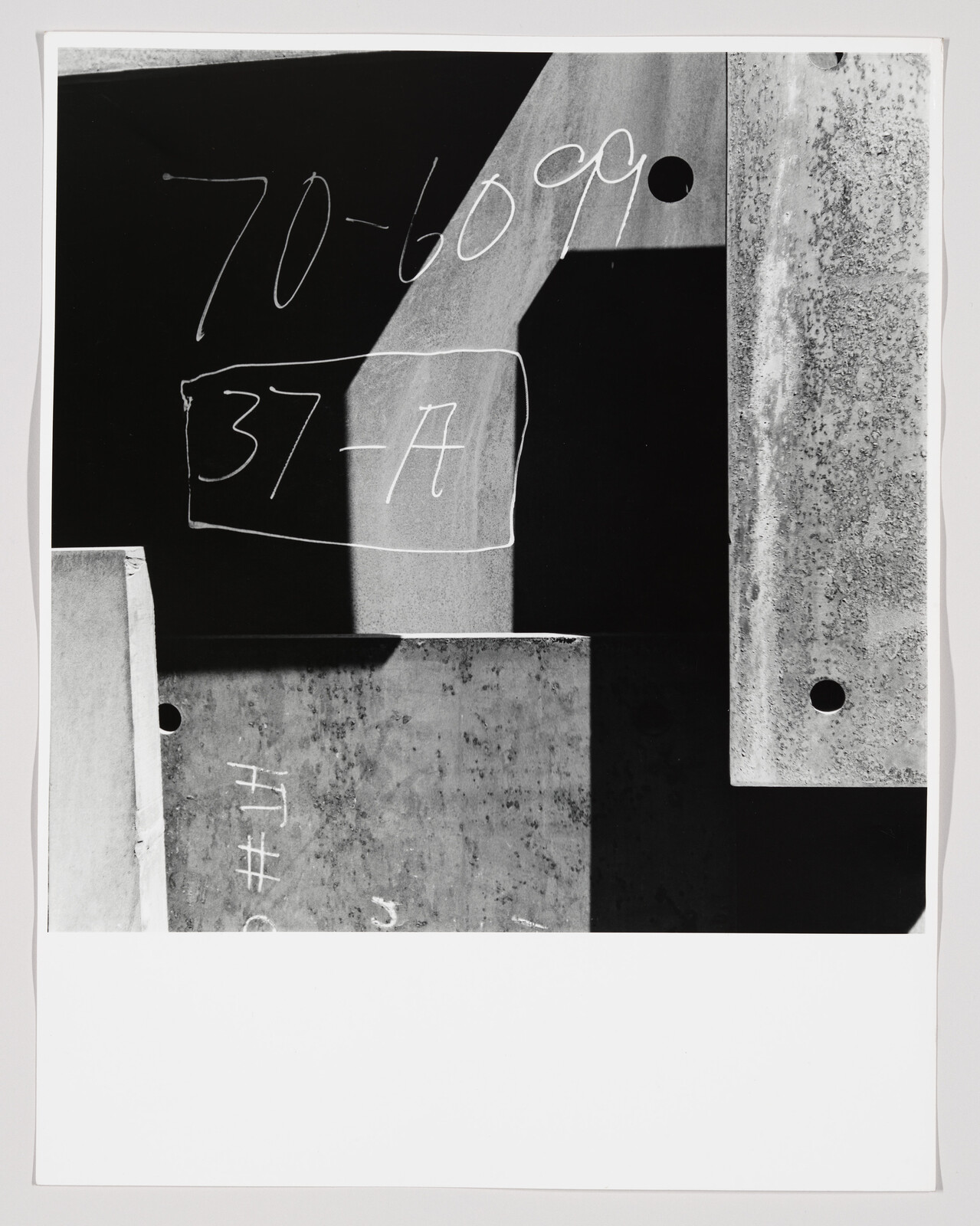 A black and white photograph featuring various geometric shapes and metallic textures with handwritten numbers and letters, such as "70-6000" and "37-A", on different parts of the surfaces.