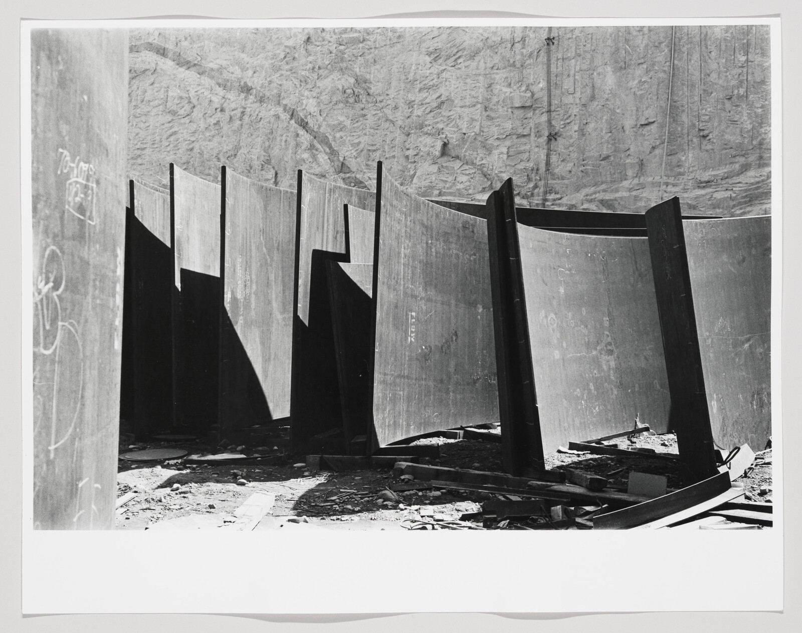 Large curved steel panels stand upright in a construction yard casting deep shadows.